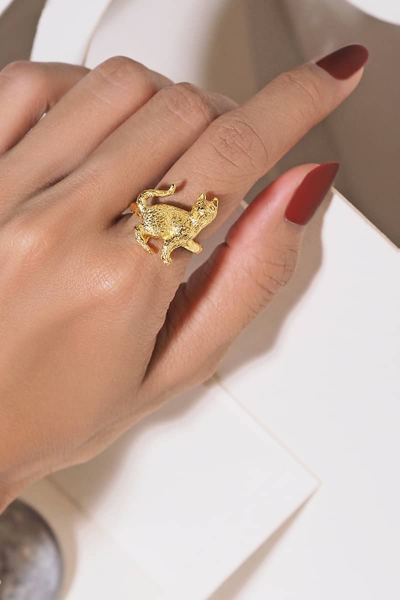 A hand with red nail polish adorned with a gold ring featuring s standing up cat design.