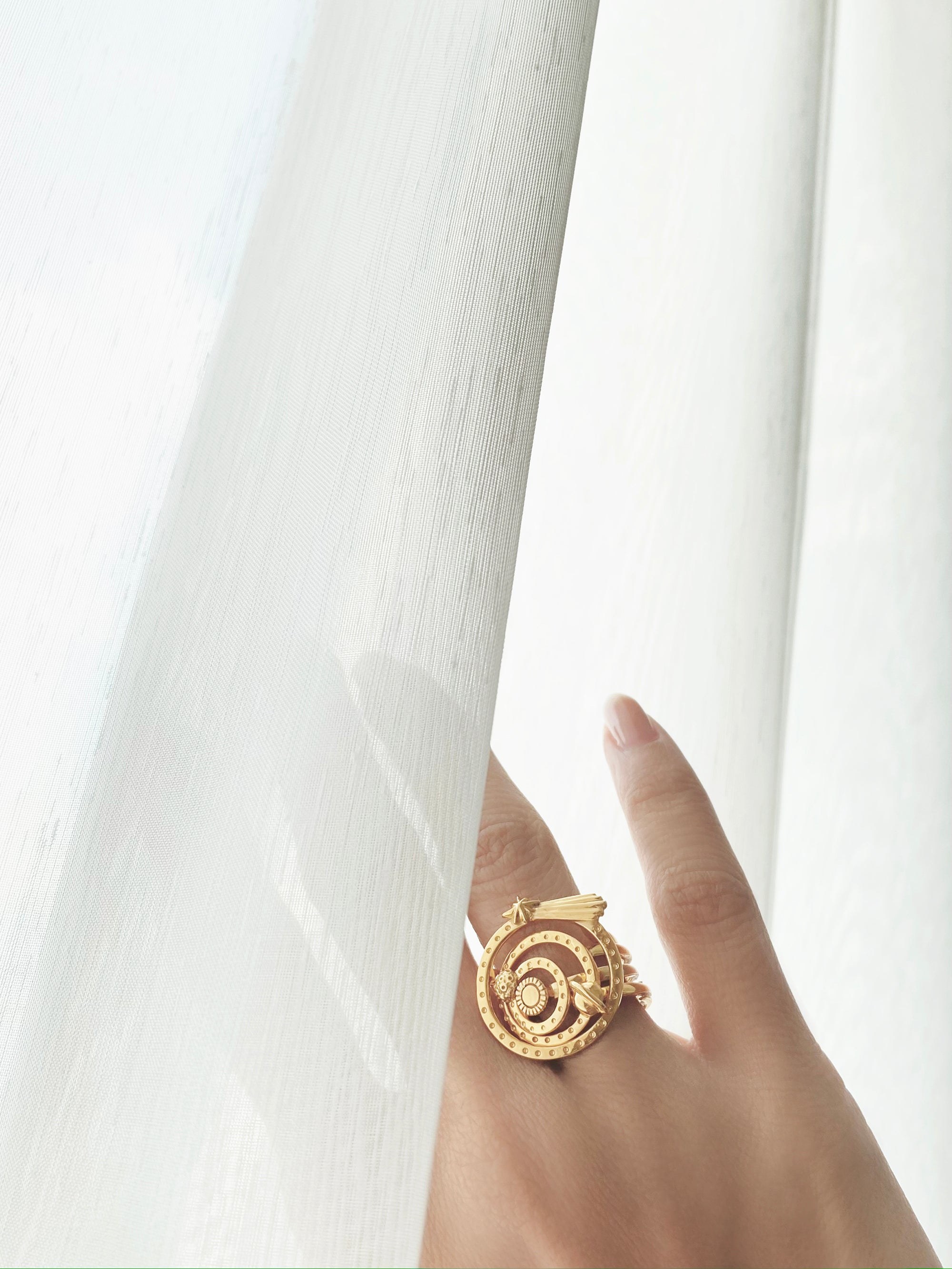 Close-up of a hand adorned with solar system-inspired gold ring set  hide behind the white curtain.