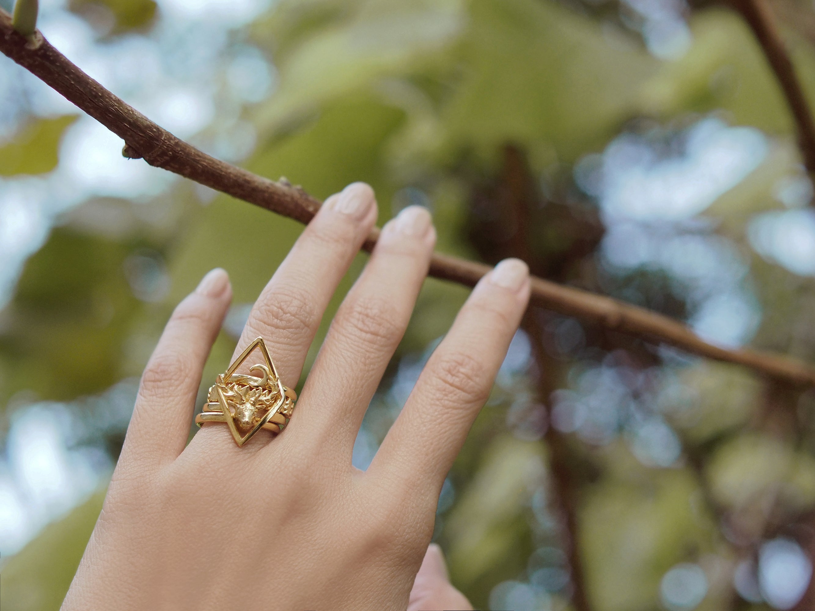 A hand wearing a deer stacking ring set, reaching for a branch in a natural setting.