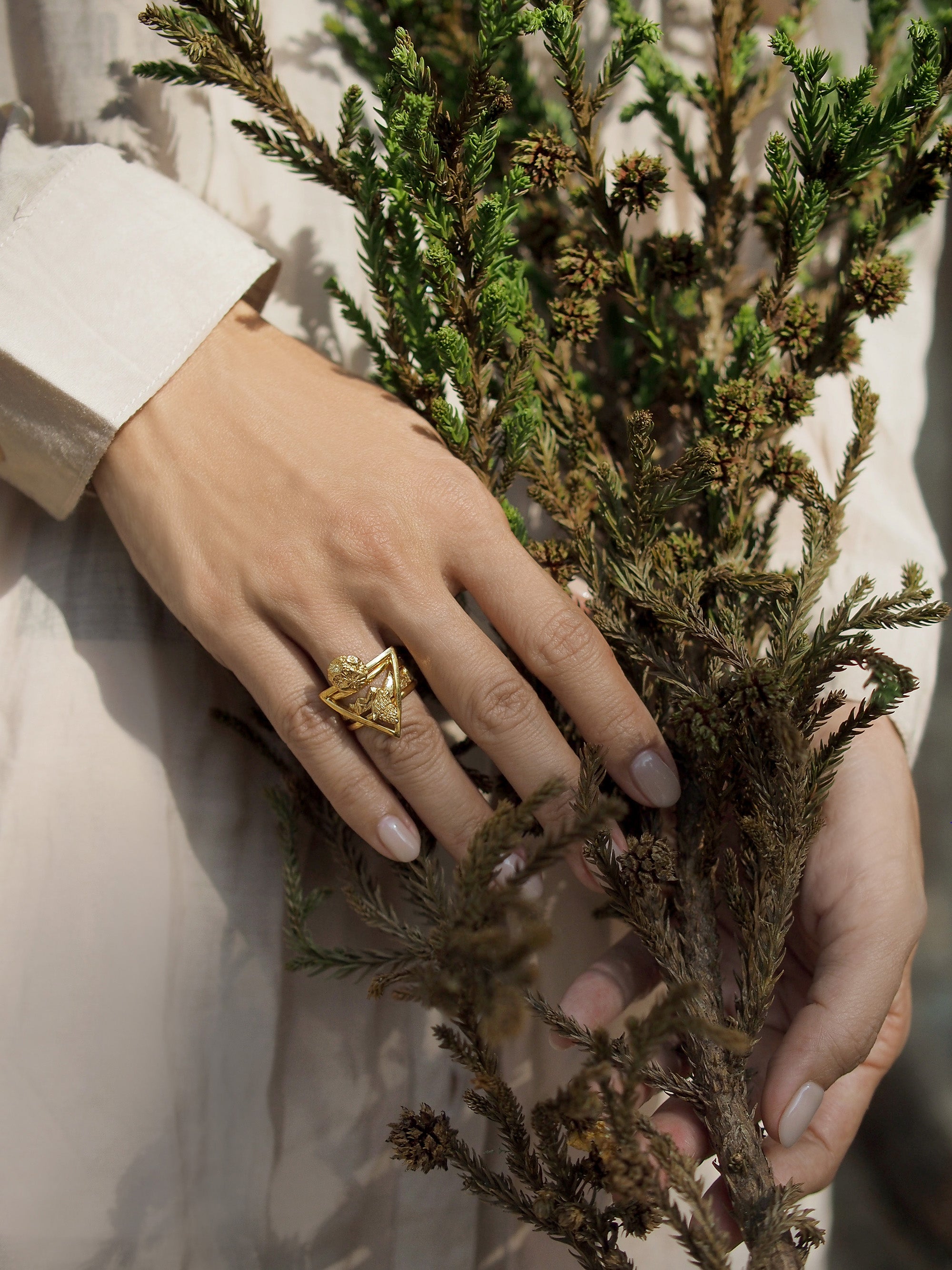 A close-up image of a hand wearing a gold wolf stacking ring set, positioned among green branches.