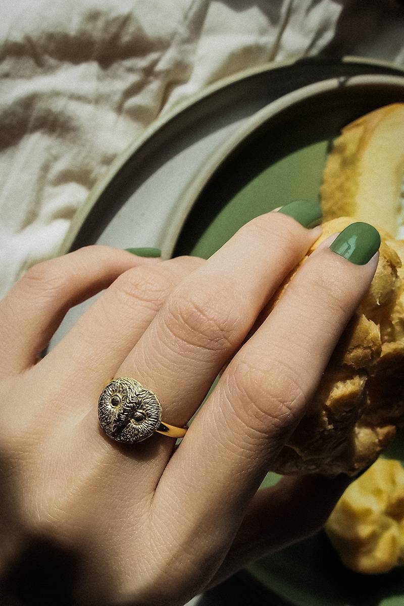 A close-up of a hand with green nail polish holding a croissant, adorned with a unique gold ring featuring an owl design.