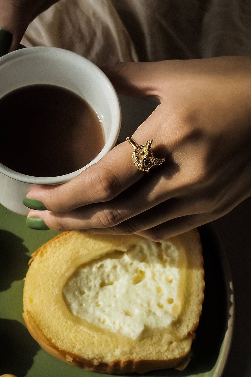 A close-up of a hand with green nail polish holding a cup of coffee, adorned with a unique gold ring featuring an owl design.