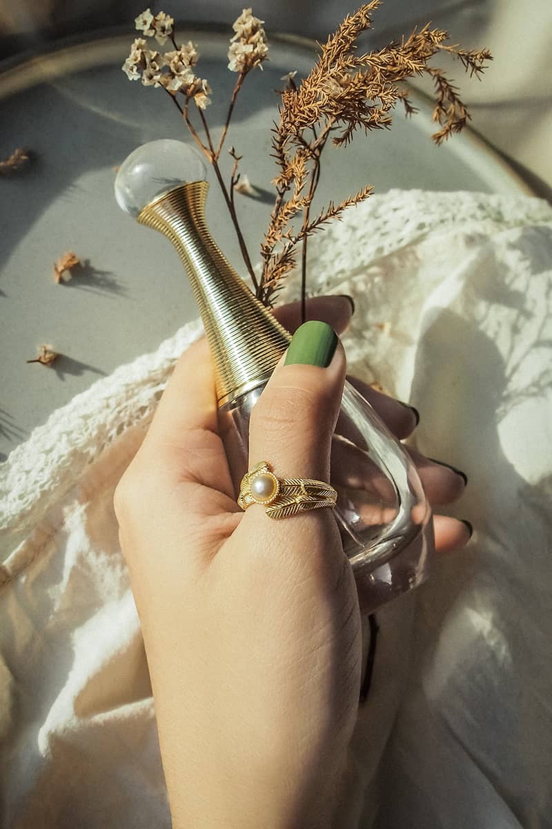 A hand with a gold ring featuring intricate designs while holding a vintage perfume bottle with dried flowers in the background.

