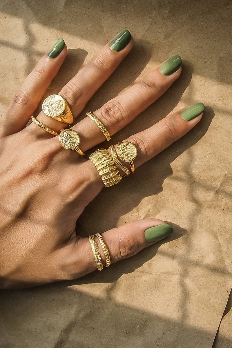 A close-up of a hand displaying multiple gold rings, featuring various designs, including forest, mountain, and owl feather designs, rests on textured brown paper.