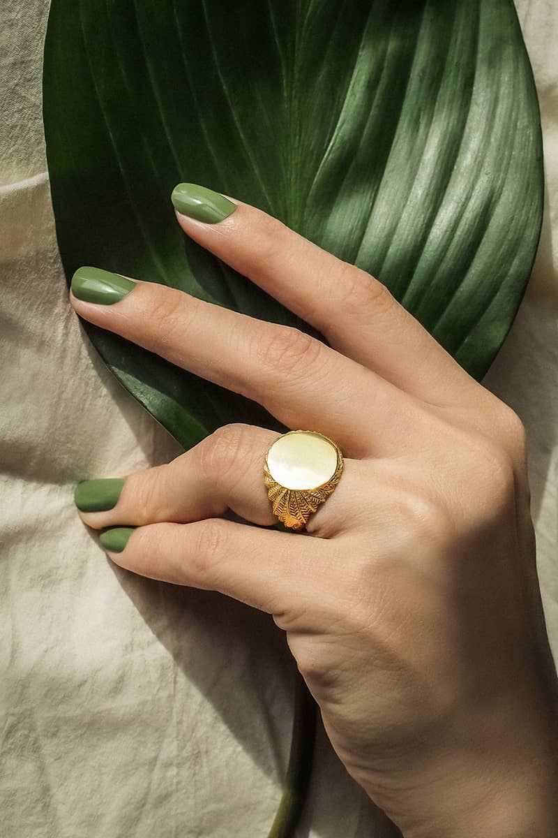 A close up of a gold ring with a smooth circular top and ornate feather-patterned band worn on a hand