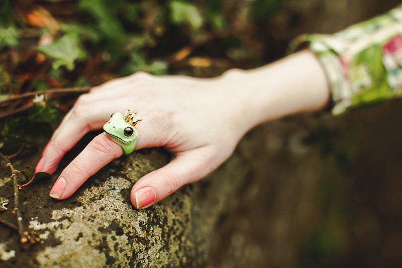 A hand wearing a frog ring with a crown, resting on a stone surface surrounded by greenery.