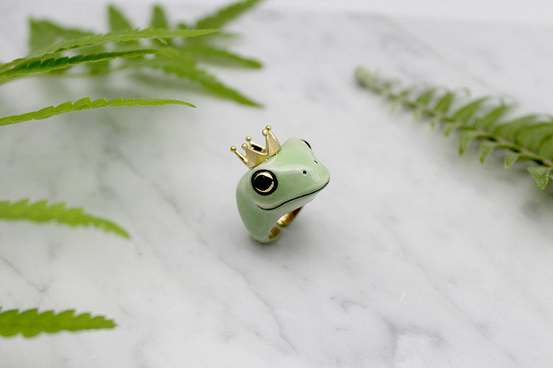 A whimsical frog ring adorned with a golden crown, surrounded by green ferns.