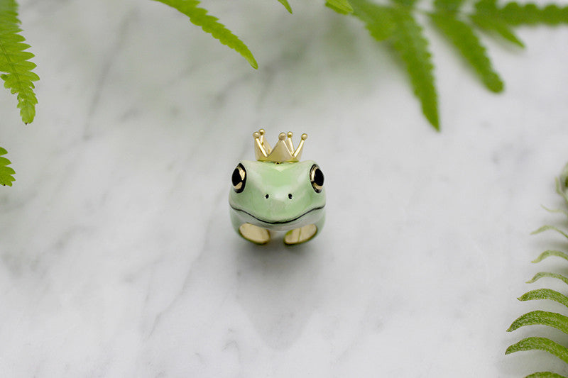 A whimsical frog ring adorned with a golden crown, surrounded by green ferns.