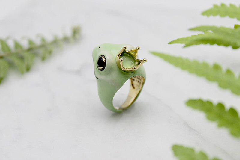 A whimsical frog ring adorned with a golden crown, surrounded by green ferns.