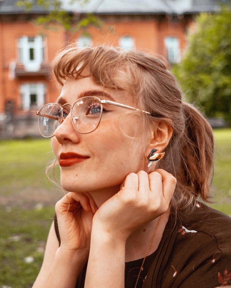 A smiling woman wearing mallard duck earrings and glasses in a park.