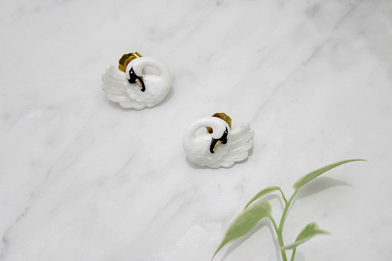 Pair of white swan-shaped earrings with black accents placed on a marble background with green leaves.