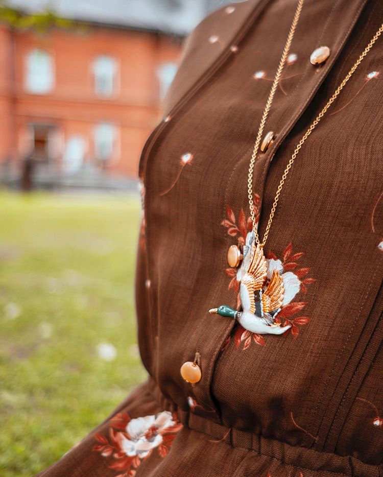 A close-up of a brown dress adorned with floral embroidery, featuring a colorful mallard duck necklace.