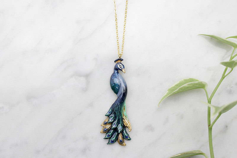 Charming peacock pendent necklace with intricate feather details placed on a marble background with green leaves.