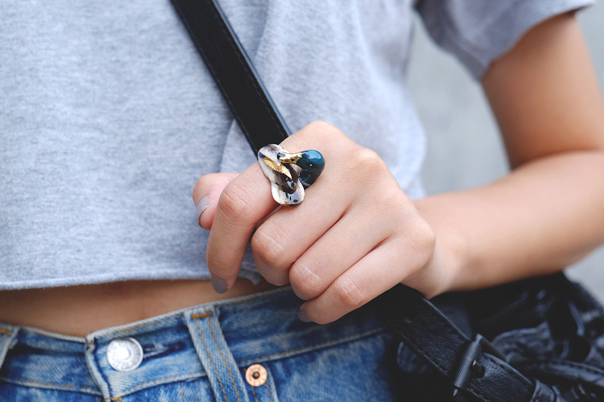 A close-up of a hand wearing a mallard duck ring with a unique design, holding a black bag, paired with a grey crop top.