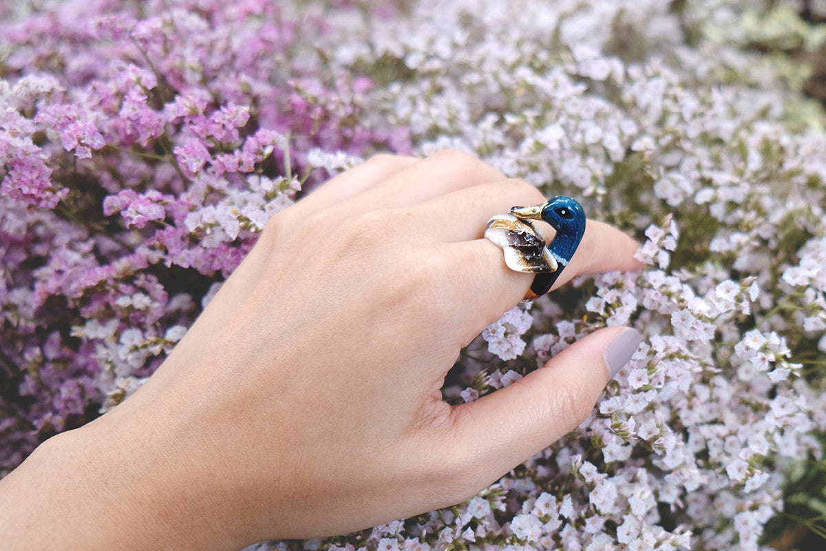 Close up of a hand wearing a mallard duck ring surrounding by flowers.