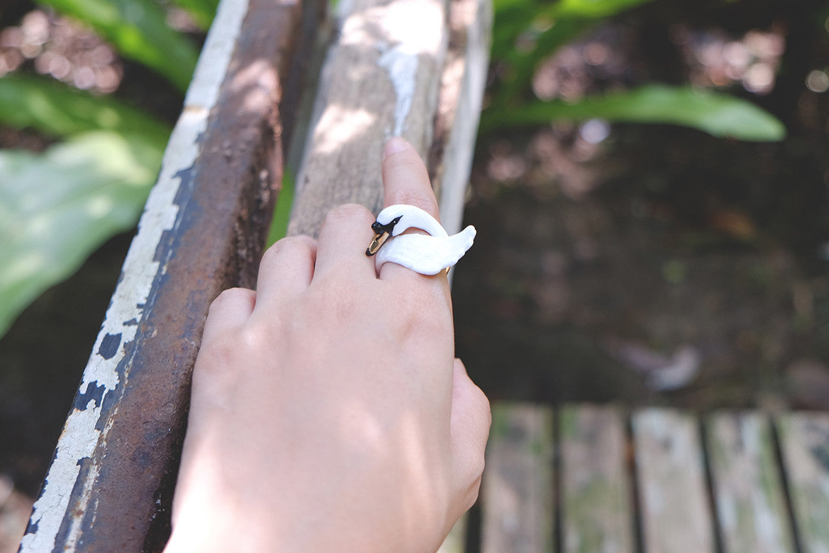 Close-up of a hand with a swan ring places on a wooden rail in a natural setting.