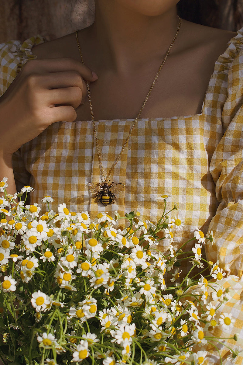 Woman wearing a bee necklace with a yellow checkered dress, surrounded by daisies.
