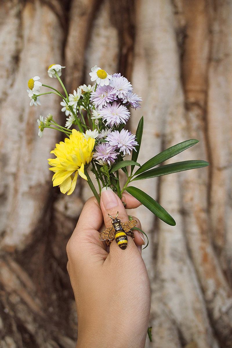 A hand holding a mix of flowers, featuring a beautiful bee ring, set against a textured tree background.