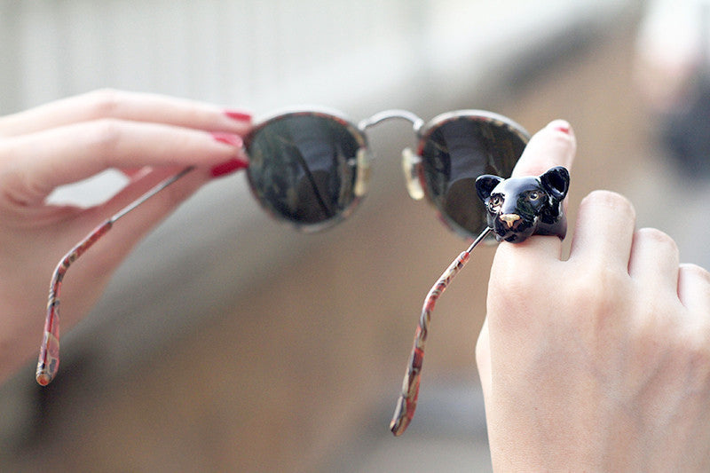 A close-up of a hand waring a black panther head ring while holding a sun glasses.