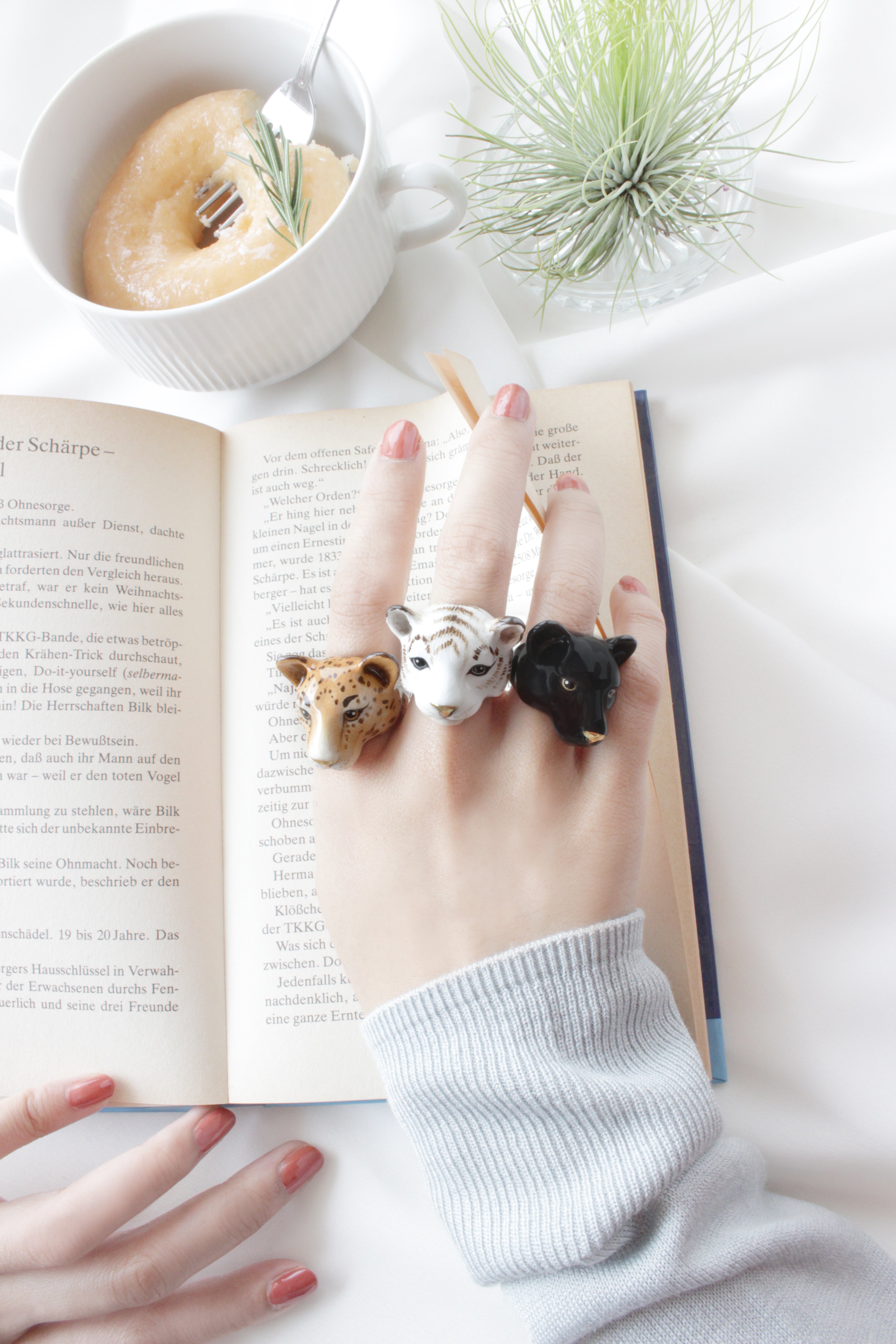 Close-up of a hand wearing animal-themed rings, showcasing a leopard, white tiger, and black panther, alongside a book and a cozy setting.