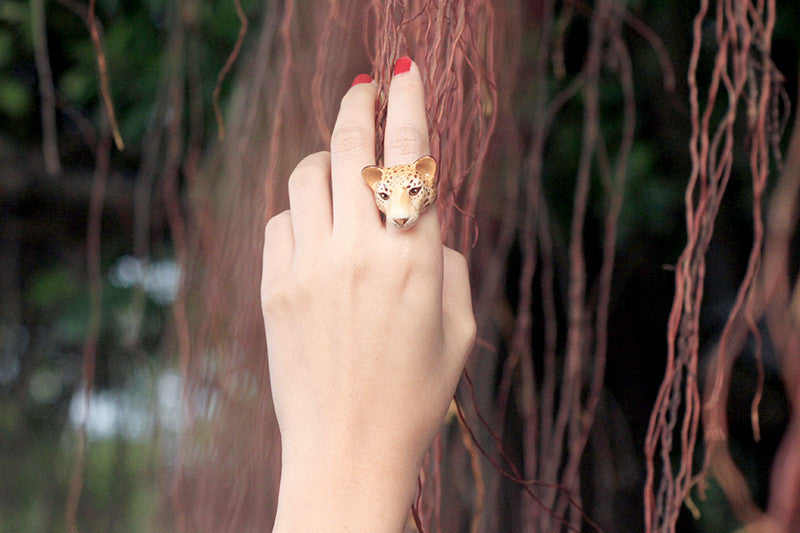 A hand with red nails displays a unique leopard ring, set against a backdrop of long, tangled vines.