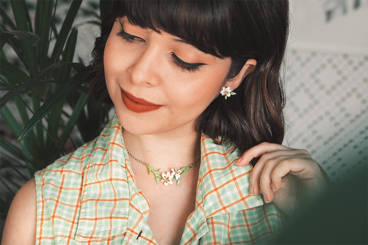 A woman wearing a checkered green blouse, adorned with floral jewelry, lily earrings and lily necklace, poses against lush greenery.