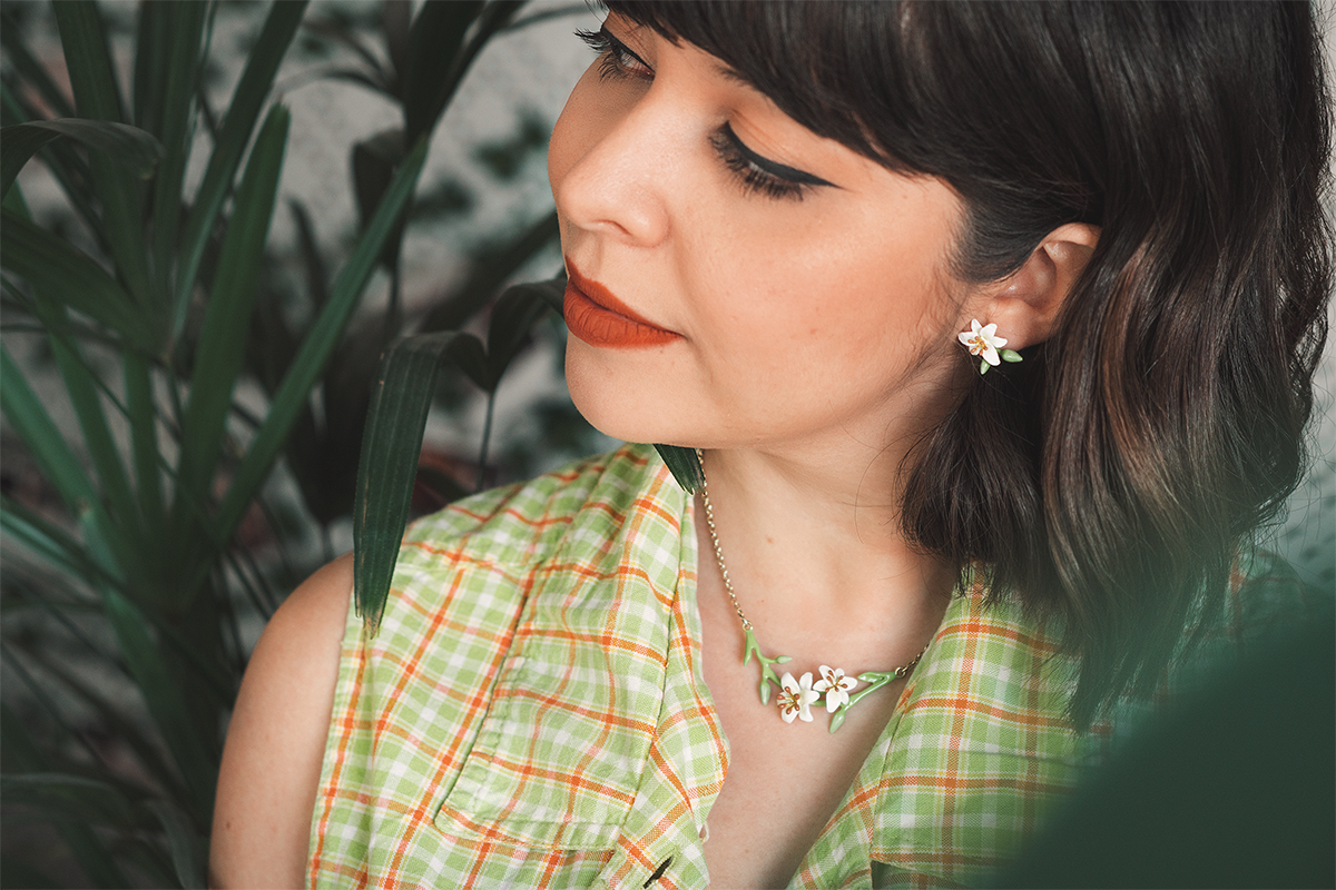 A woman wearing a checkered green blouse, adorned with floral jewelry, lily earrings and lily necklace, poses against lush greenery.