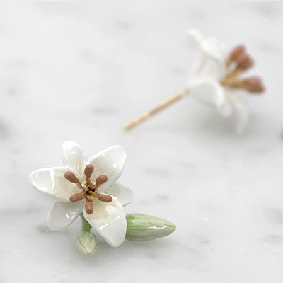 Beautiful flower earrings with white petals and brown stamens, accompanied by green leaves and a budding flower, placed on a light background.