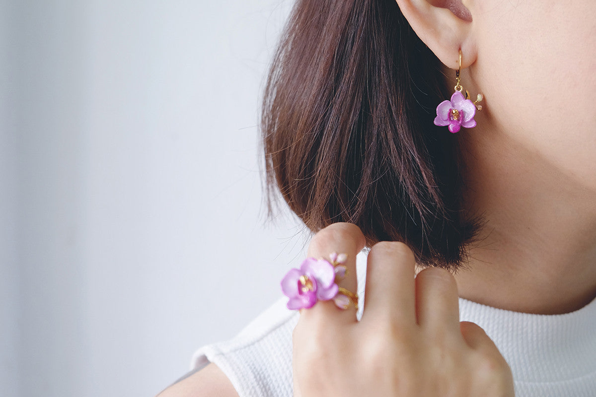 A close-up of a woman wearing floral jewelry with a purple phalaen design, including earrings and a ring.