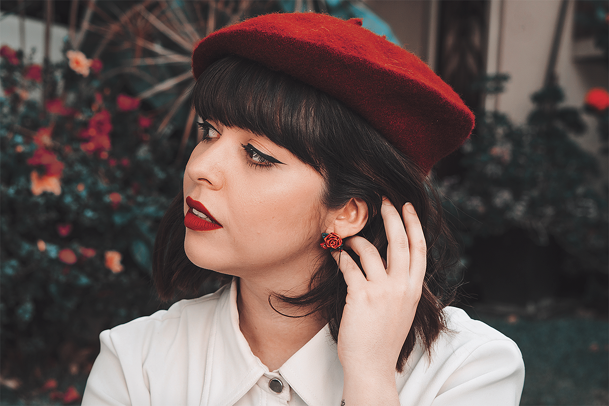 A woman wearing a red beret and a rose earring, touching her hair with a background of blooming flowers.