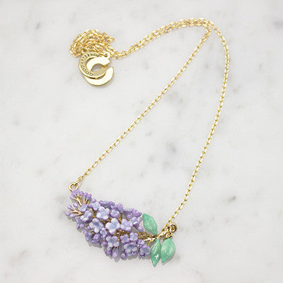 Delicate gold necklace featuring a cluster of lilac flowers and green leaves, placed on a marble background.