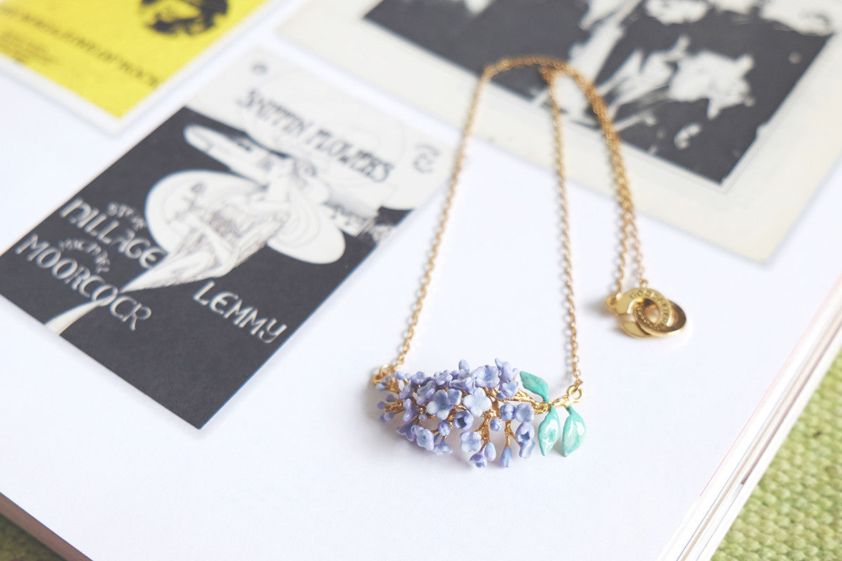 A beautiful gold necklace featuring a cluster of lilac flowers and green leaves, placed on a book.