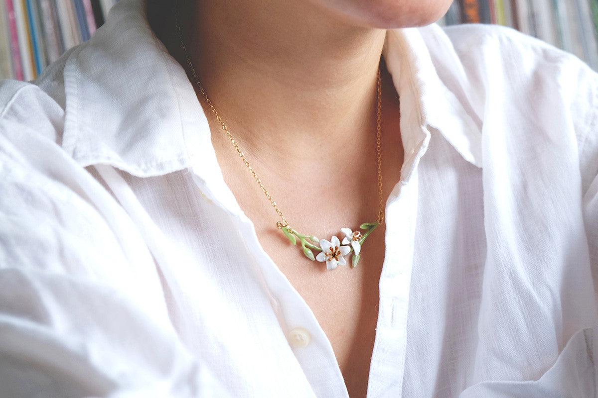 A close-up of a person's collarbone wearing a delicate gold necklace adorned with white flowers and green leaves.