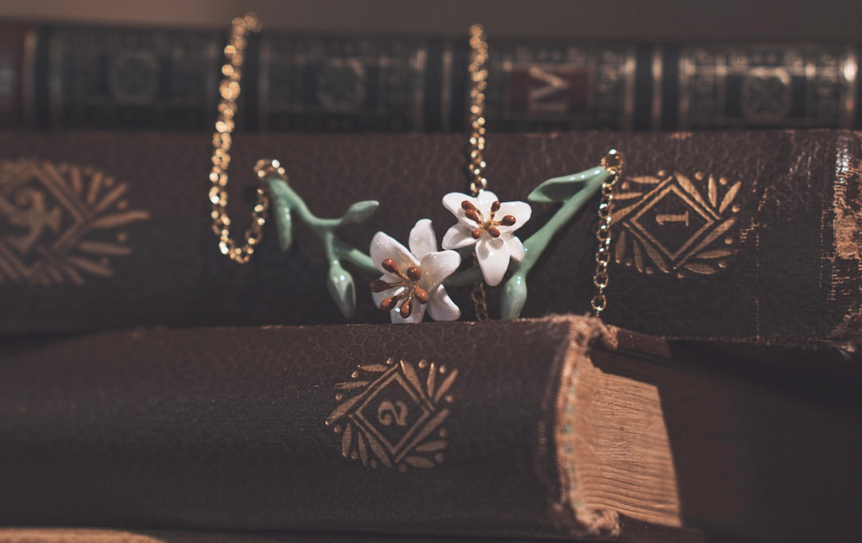 A delicate floral necklace featuring a lily design with white flowers and green leaves, placed on a pile of vintage books.