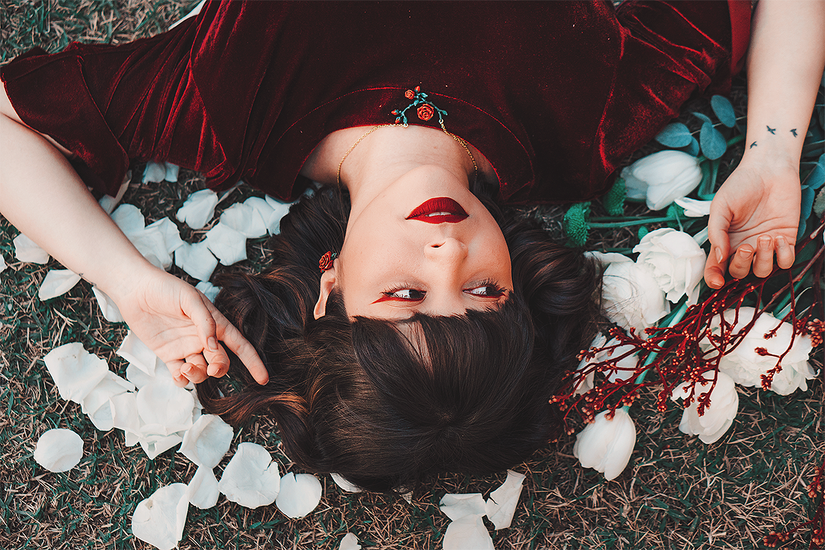 Woman lying on grass surrounded by white rose petals, wearing a rose necklace and earrings.