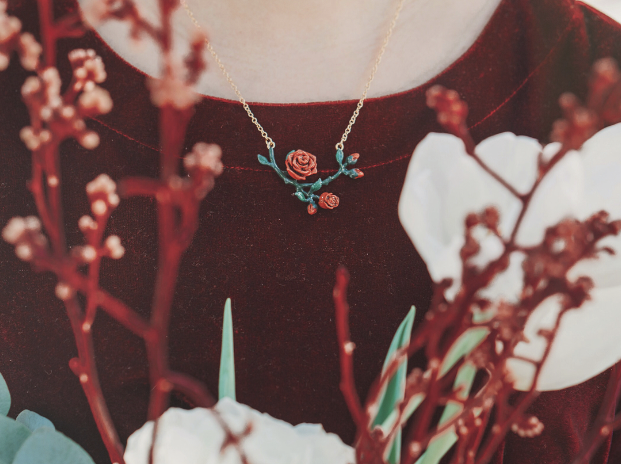 A close-up of a person in a dark burgundy garment wearing a floral necklace with red rose design, surrounded by reddish branches.