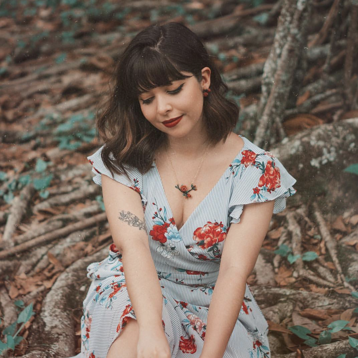 Woman wearing a floral dress, a rose necklace and earrings, sitting amidst greenery.