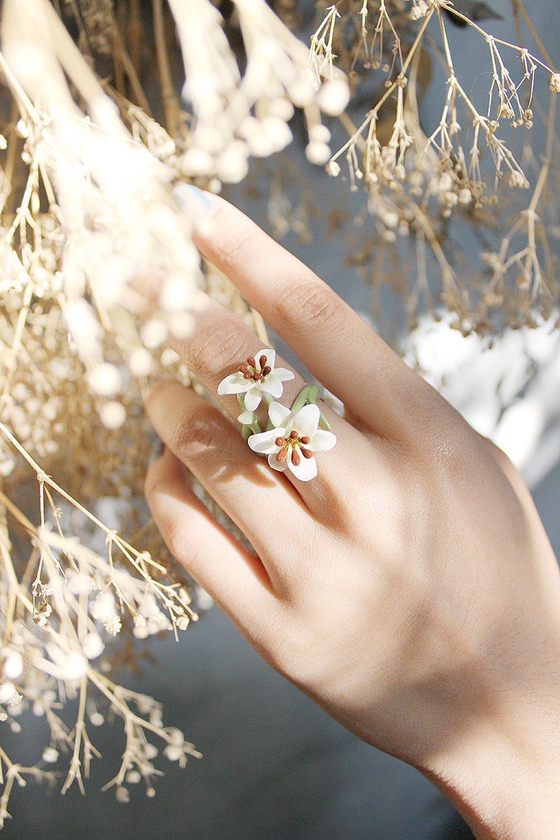 A hand adorned with a delicate floral ring featuring lily flowers design, surrounded by dried floral elements.