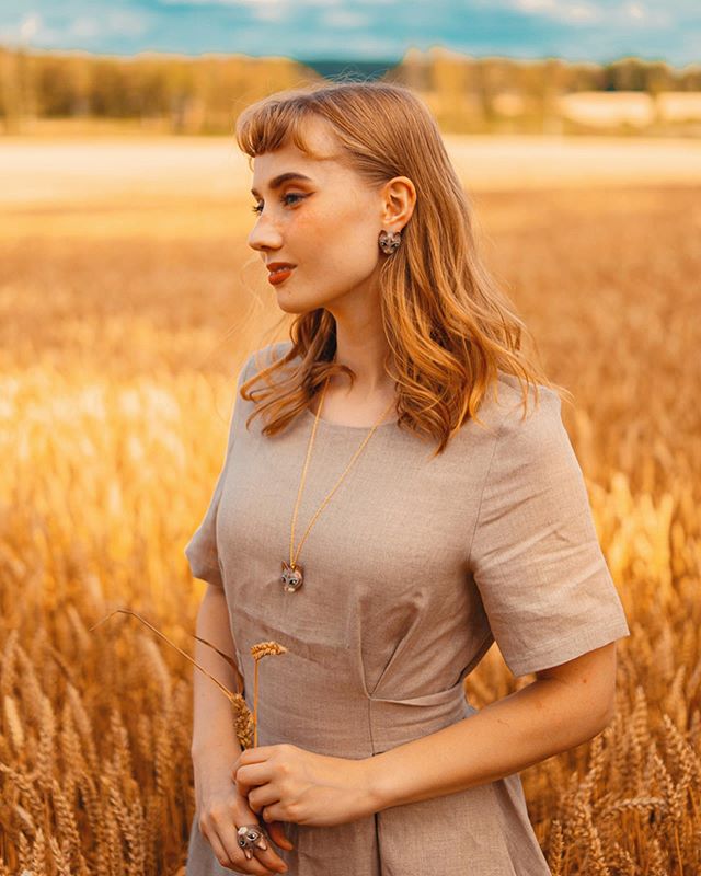 Woman wearing a cat necklace and ring, standing in a wheat field during golden hour.