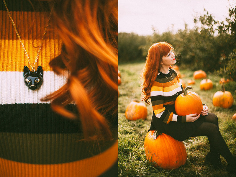 Black cat necklace worn by a person in a striped sweater among pumpkins in a field.