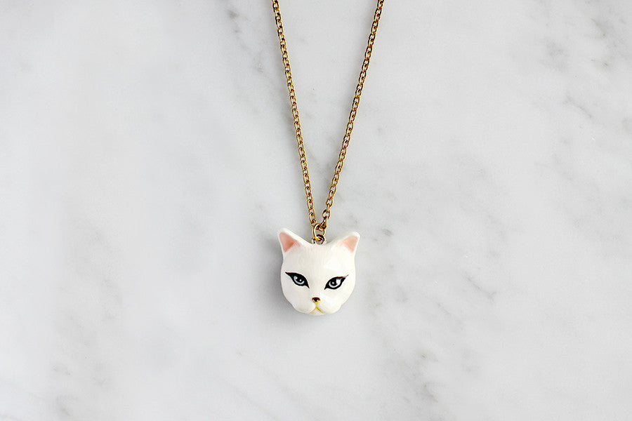 White cat pendant necklace with black eyes and pink ears on a gold chain, placed on a marble background.