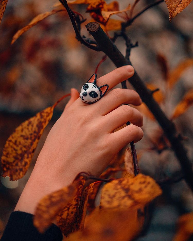 Close of a hand wearing a cat skull earrings with black and white design among the autumn leaves.