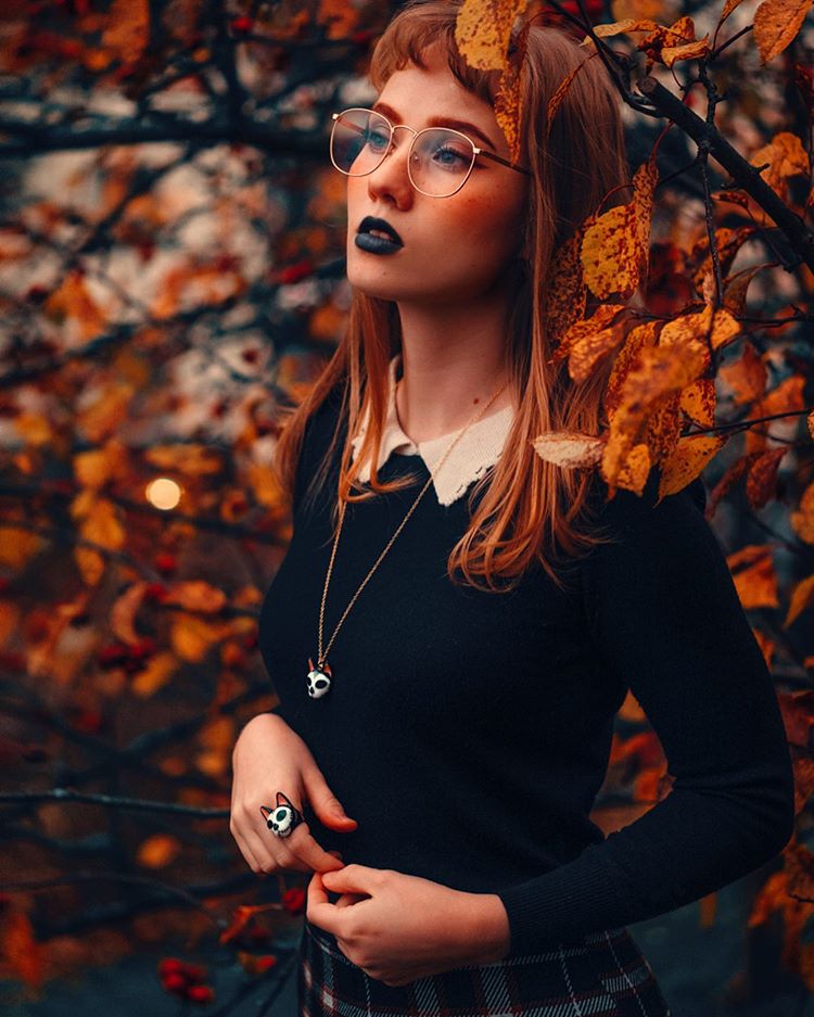 Woman with orange hair and black lipstick poses among autumn leaves, wearing glasses, a black sweater and cat skull ring and necklace.