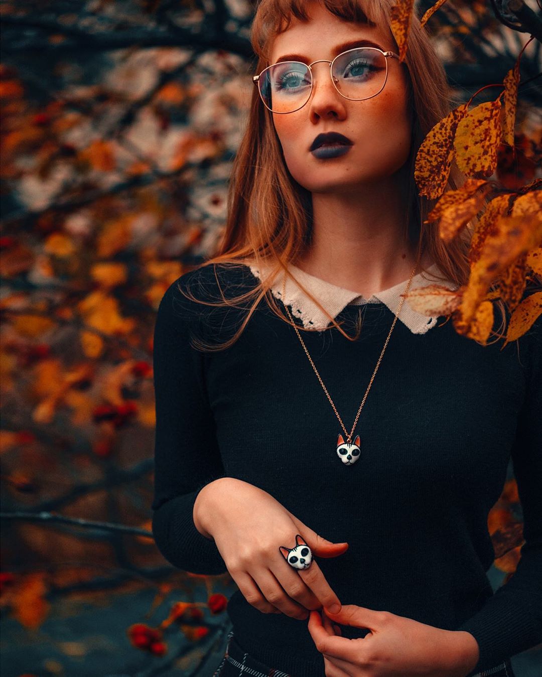 Woman with orange hair and black lipstick poses among autumn leaves, wearing glasses, a black sweater and cat skull ring and necklace.