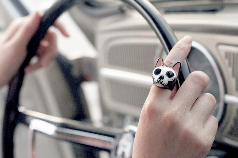 Hand holding a steering wheel, adorned with a black-and-white skull cat ring.