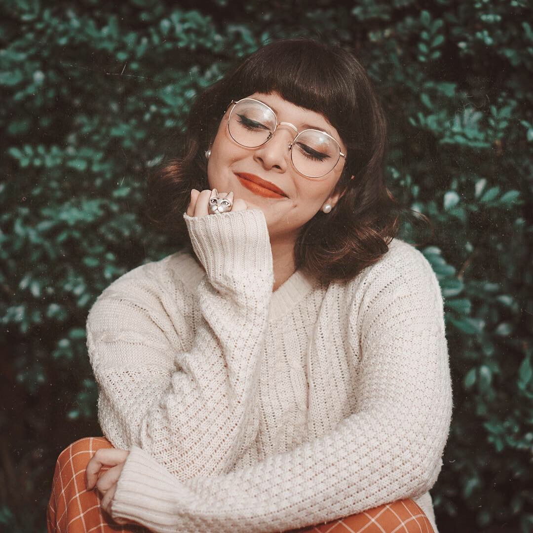 A smiling woman wearing a cat ring and a cozy sweater poses with a playful expression in front of greenery.