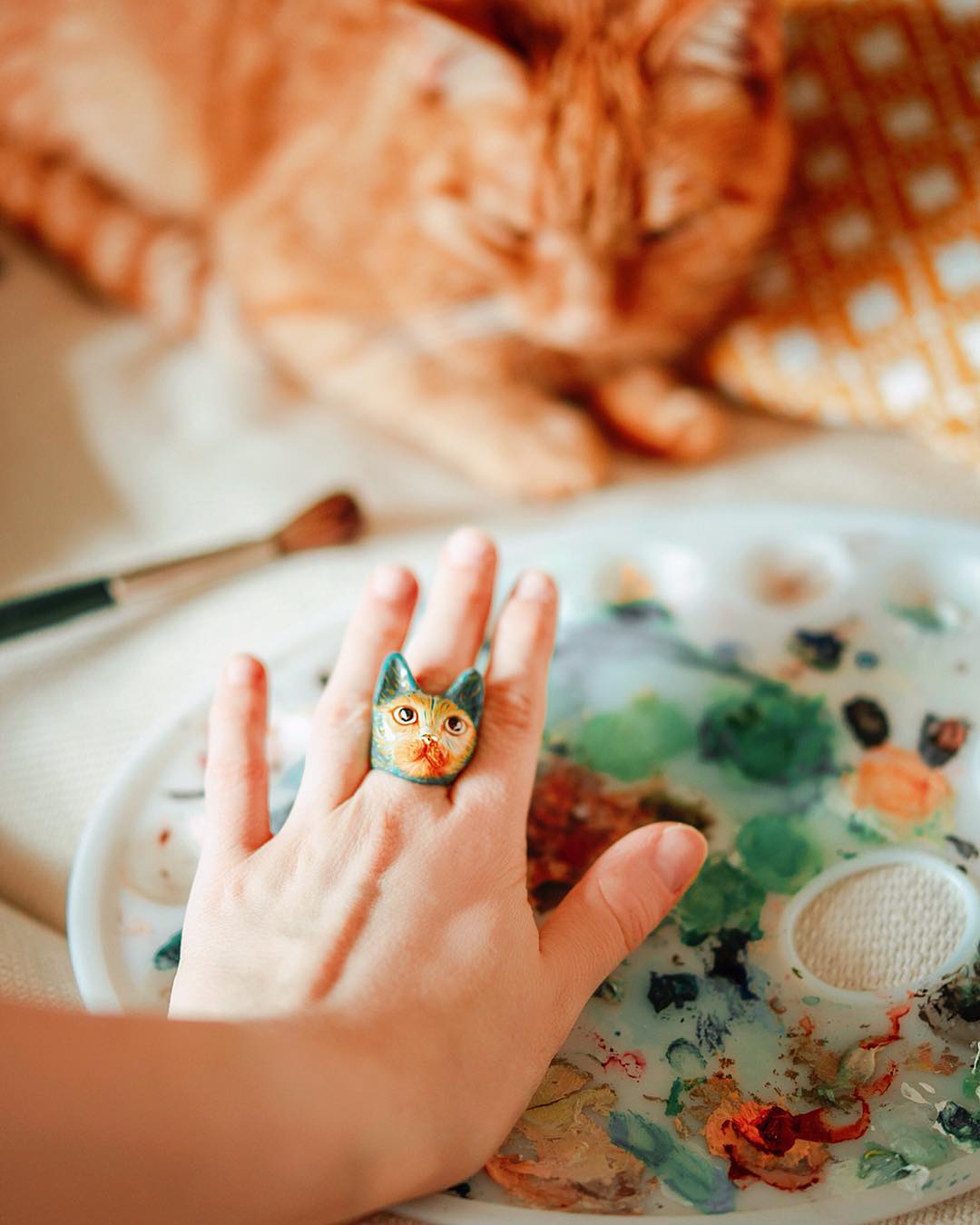 Cat-themed ring on a hand, with a paint palette and an orange cat in the background.