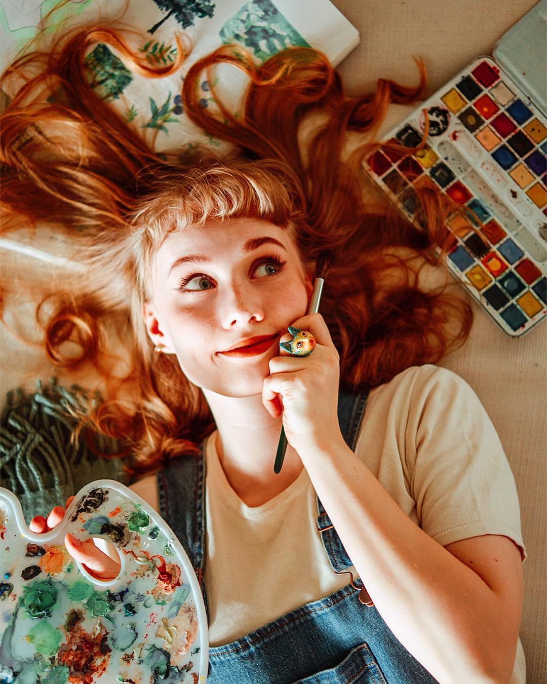 A young woman with a Vincent Van Gogh-inspired cat ring, surrounded by art supplies, smiles while holding a paintbrush to her cheek.