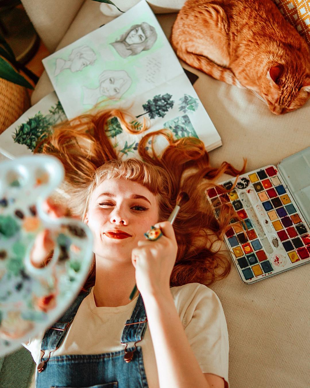 A young woman with a Vincent Van Gogh-inspired cat ring, surrounded by art supplies, smiles while holding a paintbrush to her cheek.