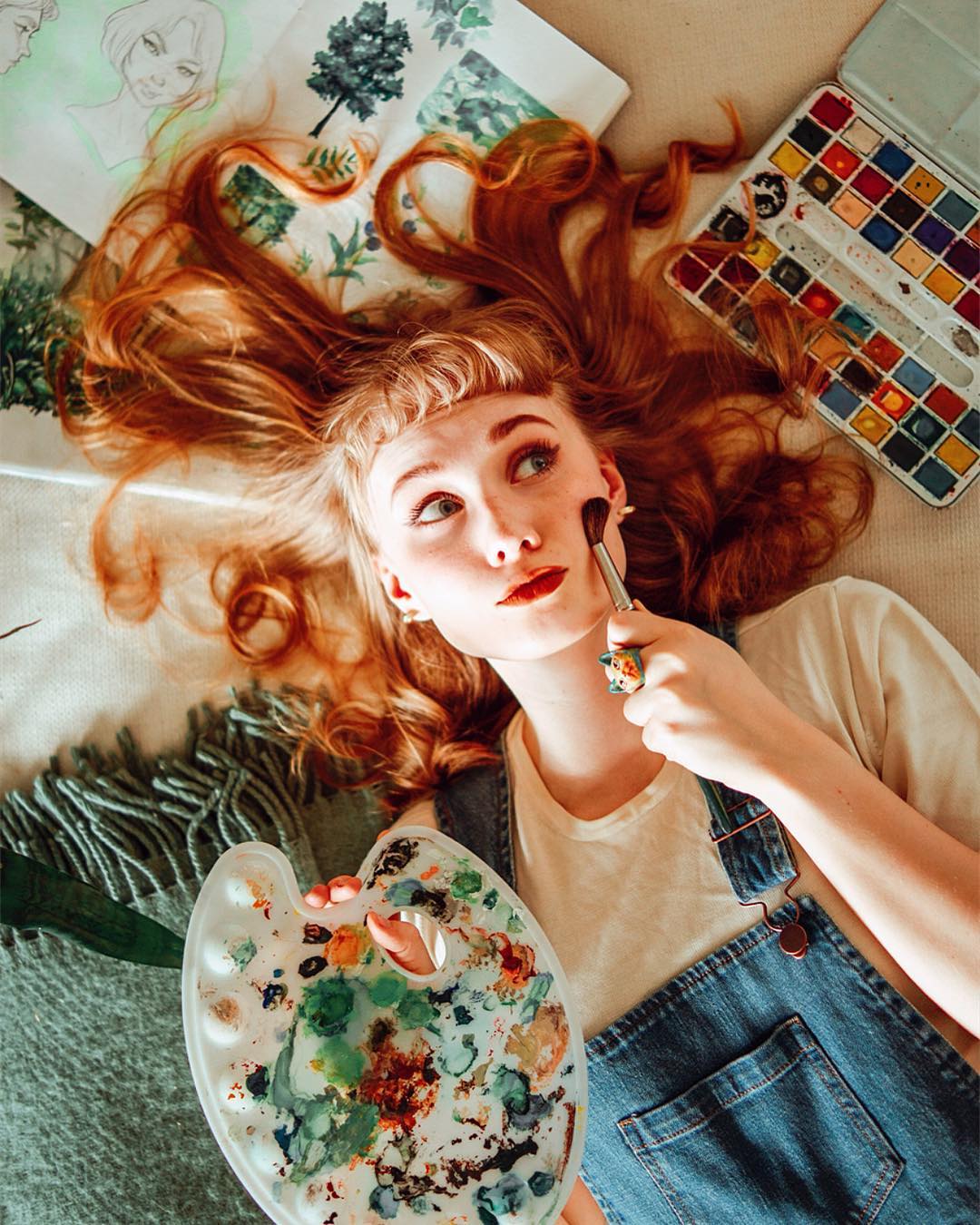 A young woman with a Vincent Van Gogh-inspired cat ring, surrounded by art supplies, smiles while holding a paintbrush to her cheek.